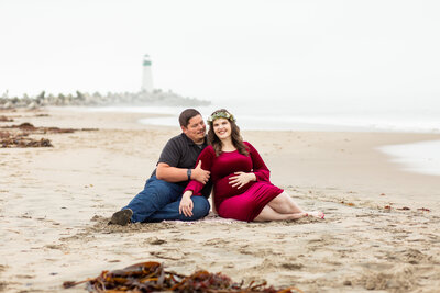 Pregnant woman and partner sit in the sand in Santa Cruz in front of a lighthouse.