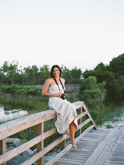 Courtney Leigh in a dress with a film camera around her neck sitting on a wooden bridge railing