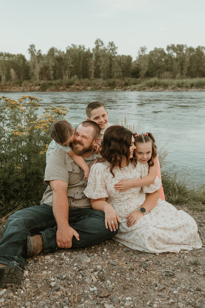 Photo of a family session taken by Bailee Freeman Photography in front of a river with yellow flowers to the left. Family is sitting on the ground and looking at each other and laughing. 