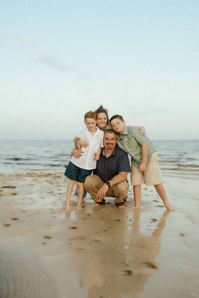 A family of four poses together on a sandy beach with the ocean in the background, smiling and embracing each other.