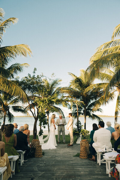A bride and groom in a cream suit stand at the altar of their boardwalk wedding under palm trees