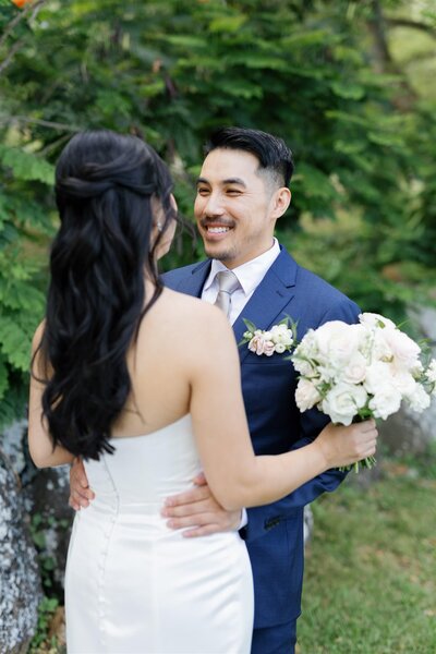 A newlywed couple with their arms around each other's waists as they smile at each other 