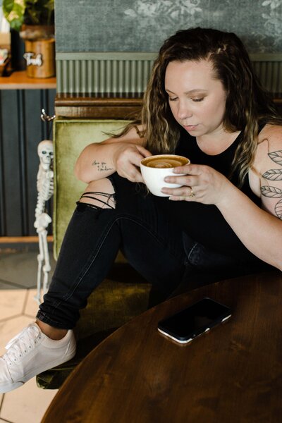 A woman with long hair and tattoos sits on a green chair, holding a large cup of coffee. Next to her phone on the wooden table, study notes for a white label web design course peek out. A small skeleton decoration is in the background.