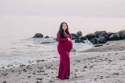 Pregnant woman in red dress smiles at camera in front of rocks at beach at Harkness Memorial State Park