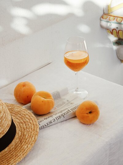 Two peaches and a glass of juice sit on top of a folded newspaper with a woven sun hat with a black band rests on the table.