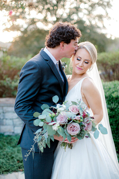 Fairytale soft pink, purple and greenery heavy bouquet featured in wedding photo of groom kissing bride on forehead