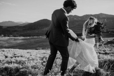 a bride and groom sitting on the ground, having a picnic with a mountain/forest view