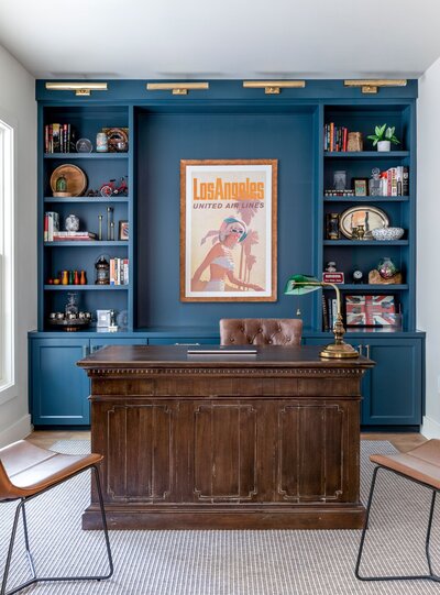navy blue office with shelves and old, vintage poster. antique desk with coper library light. carpet on wood floors. austin interior design