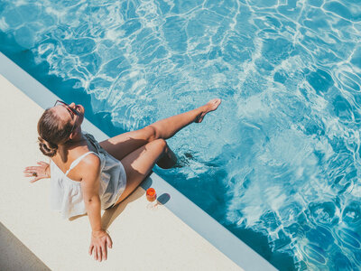 Woman relaxing by the edge of a swimming pool, dipping her feet in the water with a drink beside her on a sunny day.