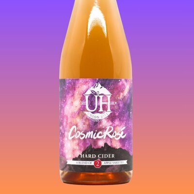 A bottle of Cosmic Rosé hard cider made by Union Hill. Label is bright pink, purple, and yellow like a nebula with Saddle Rock at the bottom of the label.