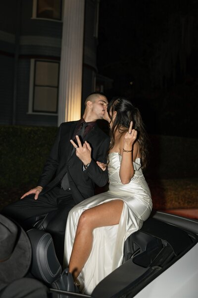 Newlywed couple sharing a kiss during their bold wedding exit in a convertible at Dr. Phillips House in Downtown Orlando, Florida — bride in satin dress, both making playful hand gestures at night.