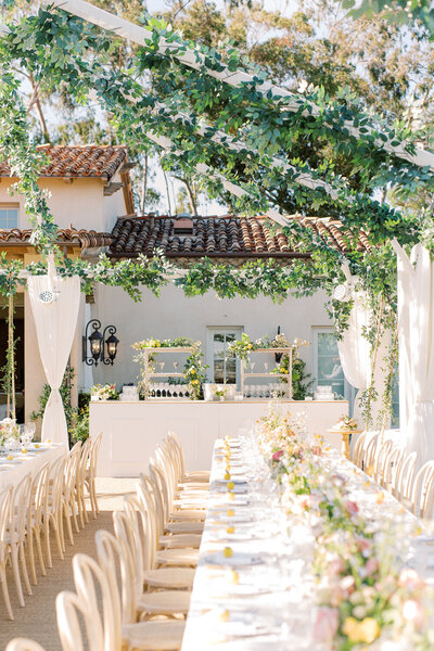 Outdoor estate wedding reception space, the building is white with red clay spanish style roof tiles, the space is decorated with long white tables, wooden chars, and plants and ivy hanging from above