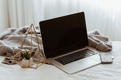 portátil abierto sobre una cama con una manta al fondo y una pequeña planta al lado