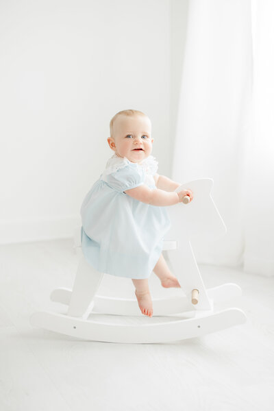 one year old baby girl sits on a white rocking horse in a blue dress during first birthday photography session