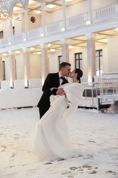 Couple posing after their wedding in a dipping style kiss surrounded by snow with the venue in the background.