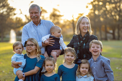 A generational family portrait in a Dallas park featuring grandparents holding their youngest grandchild, surrounded by six other grandchildren ranging from toddler to preteen. Captured in a natural outdoor setting, the image reflects connection, heritage, and joy across generations.
