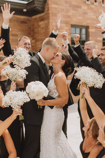Bride and groom kiss surrounded by cheering wedding party holding white bouquets at Des Moines Heritage Center wedding in Iowa.