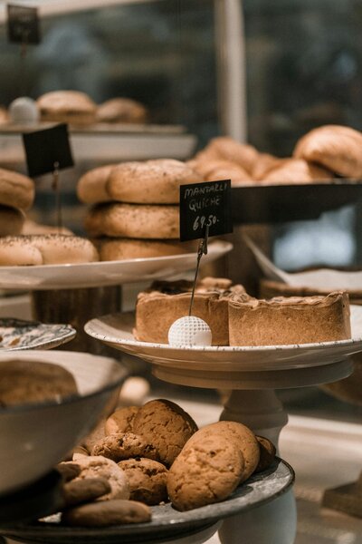 A bakery display showcasing a variety of baked goods on different cake stands, including cookies, pies, and bagels. A small chalk sign provides details about the items and prices.