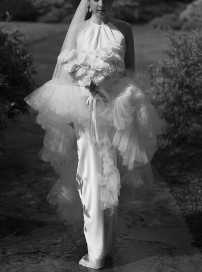 Black and white photo of a bride in a dress and a ruffled veil holding a bouquet