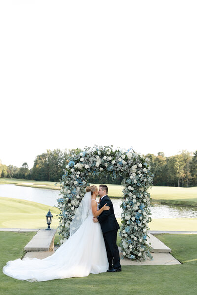 Newlyweds kiss under blue and white floral arch beside lake on golf course.