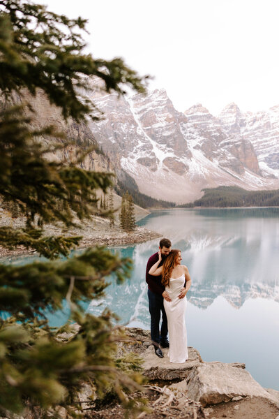 Groom, holds his bride on a rock during an engagement session Moraine lake and the mountains of Banff National Park Canada