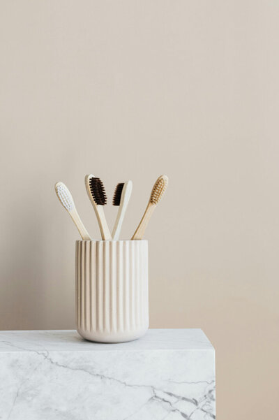 Four wooden toothbrushes in an off-white cup against a beige background, sitting on a white marble countertop.
