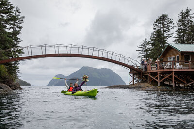 Newlyweds paddle a bright green kayak beneath a scenic wooden footbridge in coastal Alaska, with wedding guests cheering from a nearby deck. Misty mountains and ocean waters complete the backdrop of this adventurous Alaska elopement.