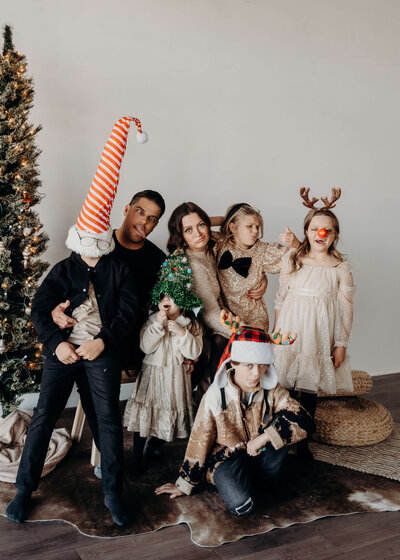 A family with five young children who is playfully next to a Christmas tree, wearing all sorts of Christmas props, like a tall, elf hat, reindeer, antlers, and a Christmas tree while smiling with their parents.
