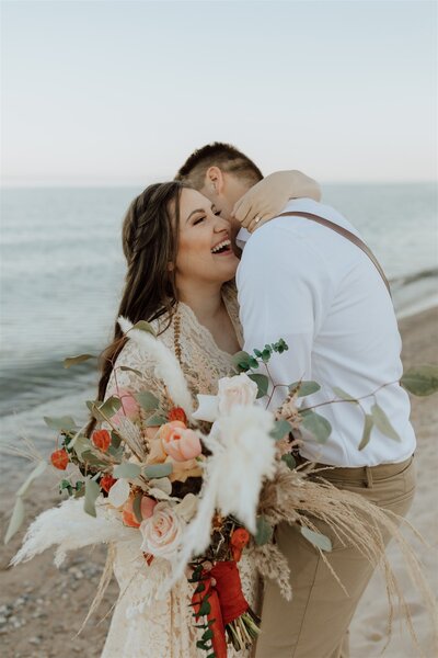 groom whispering in brides ear on a beach, bride is laughing on a beach in Wisconsin