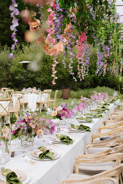 Head wedding table with dinner ware white linen light wood chairs and cascading purple and pink flowers above