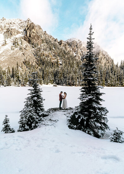 perfectly centered between two snowy pines, a couple, in their wedding attire, share vows. The mountains frame their background and they pop against the snowy landscape.