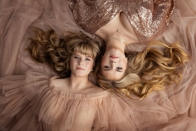 Twin sisters lying side by side on flowing blush tulle gowns with soft waves in their hair during an elegant Dallas senior portrait session.