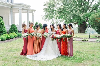 A bride and her bridesmaids, wearing verona sunset colored dresses, laugh with one another outside of John Joseph Inn. 