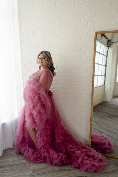 Pregnant woman in a flowing pink tulle gown posing by a window during a studio maternity session — photographed by Alliston and Barrie maternity, newborn, and family photographer.