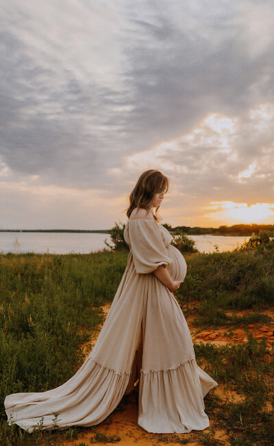 A mom standing in a field next to Grapevine Lake in Flower Mound, Texas wearing a cream dress and holding her pregnant belly 