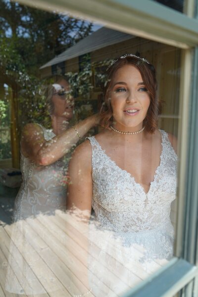 Weddings and Family Portraits in Orange County bride looking out window while mother of the bride puts on her necklace
