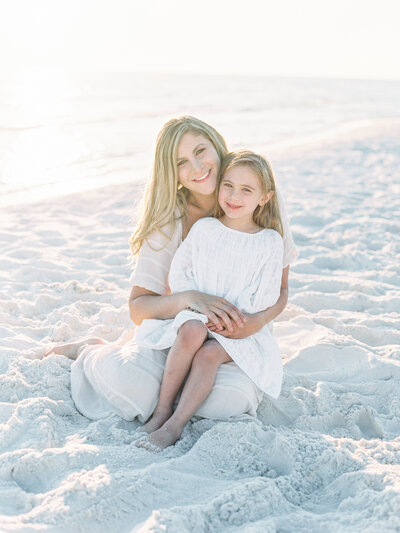 A mom and daughter smile at the camera while sitting on the beach in Santa Rosa Florida
