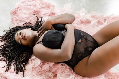 Dark skinned woman wearing black lace bodysuit posed laying on pink ostrich feathers