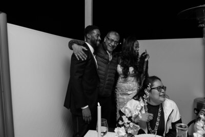 Wedding guest wrapping arms around bride and groom during the reception, in black and white.