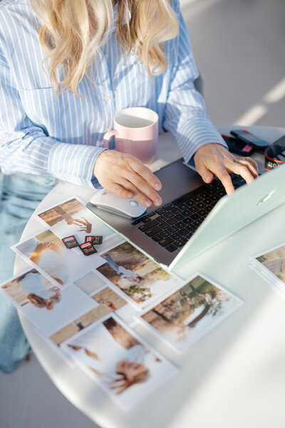 a woman types at her computer while holding a cup of coffee and is surrounded my printed photography images at the the white table she is sitting at