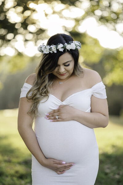 Woman wearing a white gown and flower crown looks down at her pregnant belly. Atlanta Maternity Photography by Amanda Touchstone