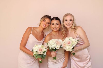 Three bridesmaids in white dresses holding bouquets, posing in front of a soft blush pink backdrop.