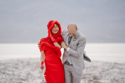 A woman in a red dress and a man in a gray suit are laughing as wind blows their clothes