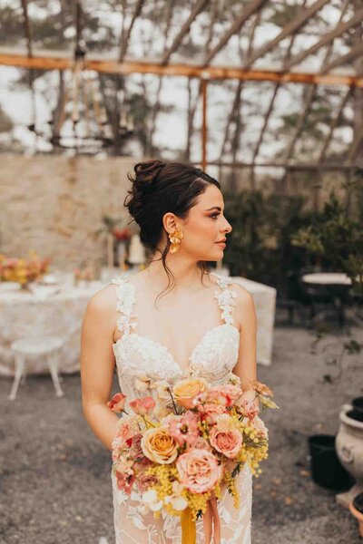 beautiful bride leaning against a column wearing her gown and holding her bouquet, under a canopy of trees.