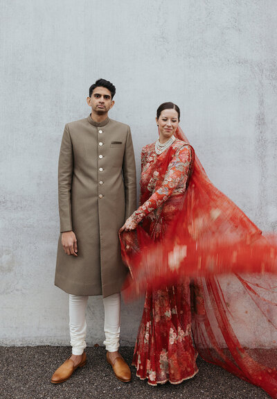 Bride in intricate red saree swishes fabric while smiling and posing with groom