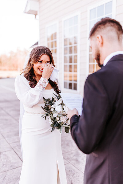Bride  holding bouquet, while crying with tissue gently to eye while groom reads his vows.