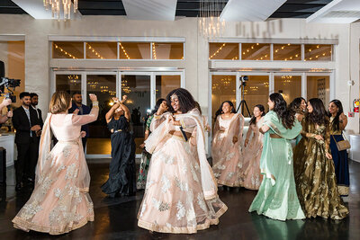 Guests in pastel lehengas dance joyfully on the black-tiled floor at SoHo63, a Chandler wedding and event venue. Surrounded by chandeliers and reflective windows, the celebration brings vibrant energy and cultural beauty to the modern indoor reception space.