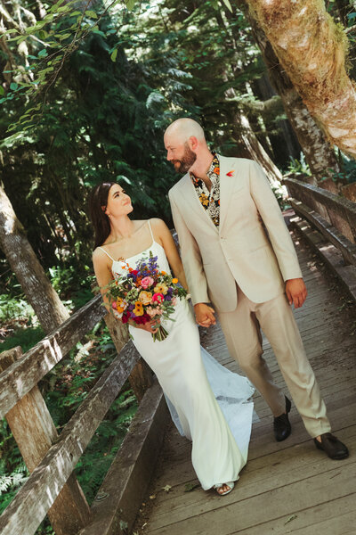 Colorful bride and groom walking along a wooden bridge holding hands and colorful bouquet