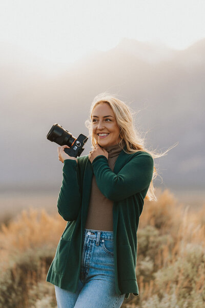 woman standing outside with mountains in the distance as she smiles while holding a camera