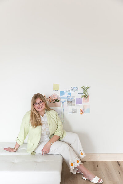 brand photography of blonde girl with glasses wearing green blazer sitting on white couch in front of inspirational mood board on wall